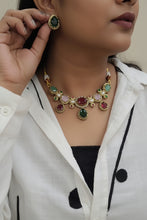 Load image into Gallery viewer, A close-up of a person wearing a gold-tone statement necklace with colorful gemstones, while holding a matching green gemstone studs between their fingers.